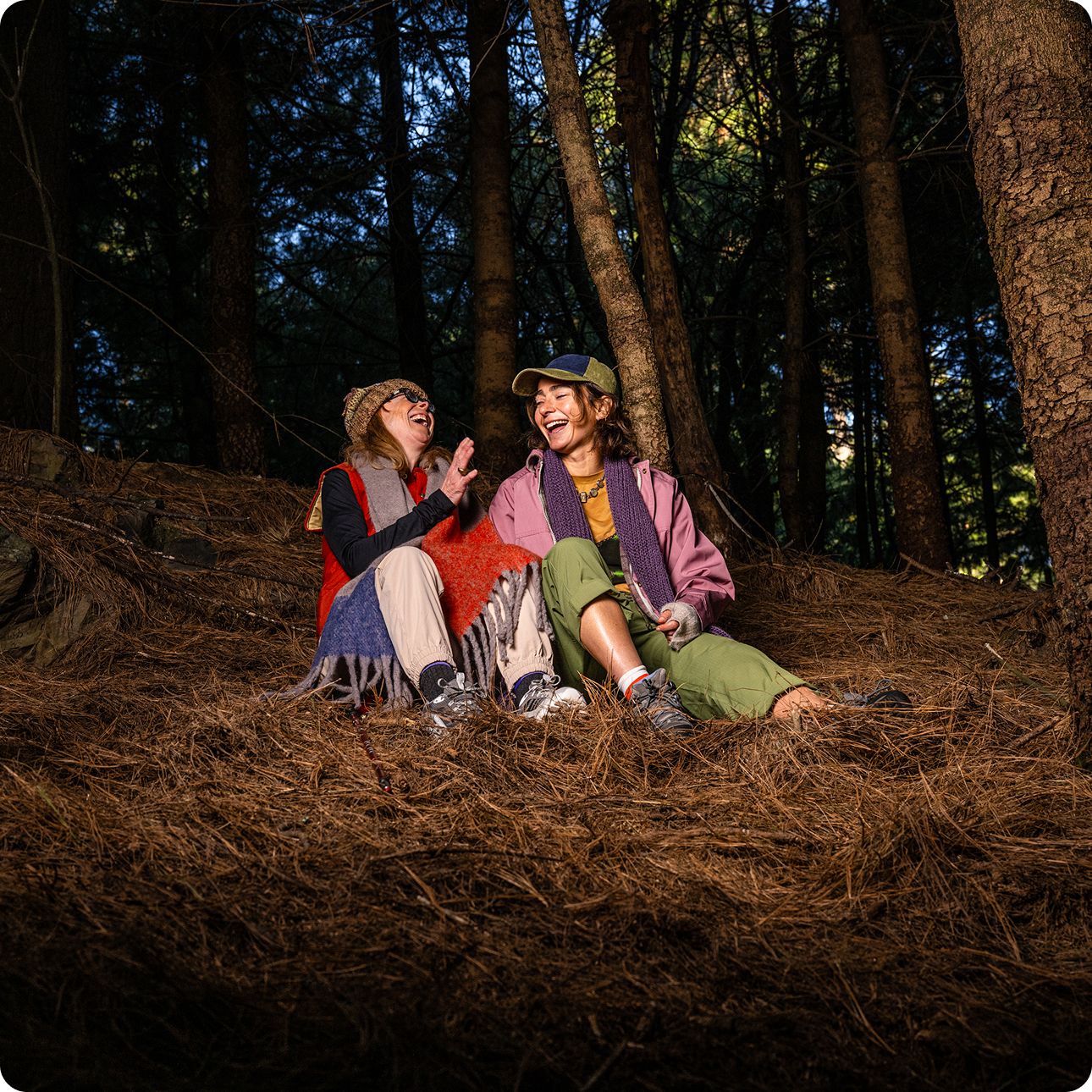 Two people in the woods, sitting together on a pile of brush, talking and laughing.
